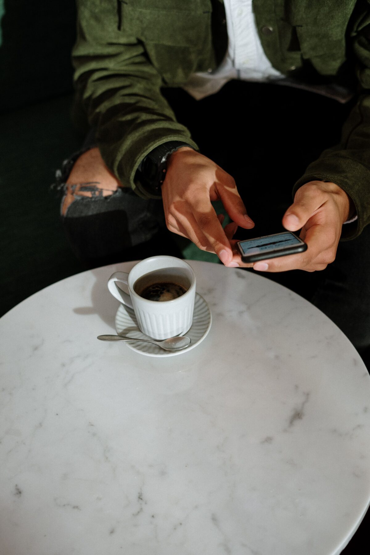 A person wearing a green jacket sits at a white marble table, using a smartphone. On the table is a cup of black coffee on a saucer with a spoon. Only the persons hands and part of their torso are visible.